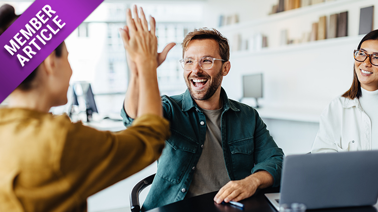 Stock image of a smiling white man in glasses and a denim shirt sitting at his computer desk giving an off-screen female co-worker a high five. Another female coworker dressed in white and wearing glasses smiles beside him.